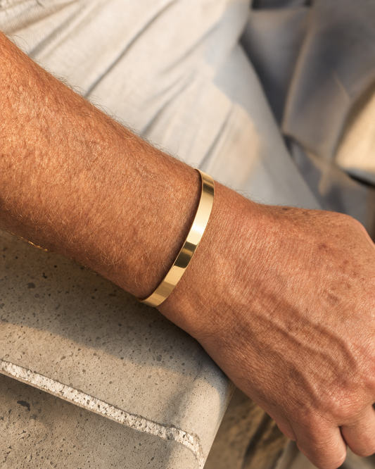 887Seven Meridian Cuff in 18K gold, 6mm flat band, worn on a man's wrist resting on a concrete ledge